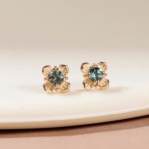 May include: A pair of gold stud earrings featuring a round, faceted blue-green gemstone at the center. Each stone is held by four prongs within a decorative, petal-like gold setting that radiates outward, creating a small, flower-inspired design.