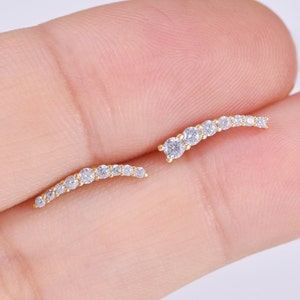 May include: A pair of gold-toned ear climbers featuring a curved design set with seven small, clear, round-cut stones. The earrings follow a gentle arc shape, with each stone held in a metal setting, creating a delicate and linear appearance.