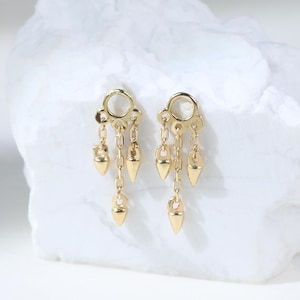 Può includere: Un paio di orecchini pendenti in oro caratterizzati da un cerchio superiore collegato a tre delicate catenine di diversa lunghezza, ognuna terminante con un piccolo ciondolo a punta in oro lucido su una superficie bianca.