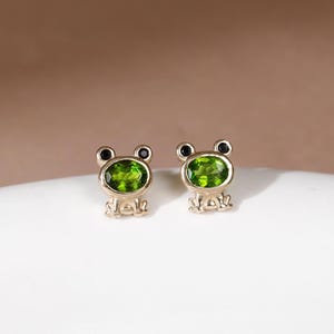 May include: A pair of gold stud earrings shaped like frogs, each featuring a small oval-cut green gemstone as the body and two tiny black circular stones for the eyes. The metal has a polished finish, and the design includes small feet at the base.