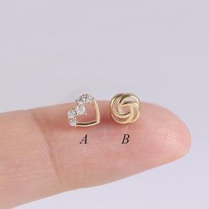 May include: Two small gold earrings. One is heart-shaped with three clear stones, and the other is a gold knot design. The earrings are labeled A and B.