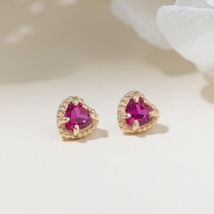 May include: A pair of gold stud earrings featuring heart-shaped fuchsia pink gemstones. Each stone is set in a gold frame with a delicate beaded edge, creating a small, detailed design that highlights the vibrant color of the stones.