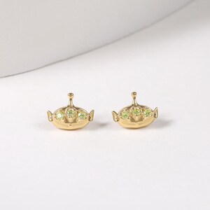 May include: A pair of gold-toned stud earrings shaped like three-eyed alien characters. Each earring features a smooth metallic finish with three small, round, light green gemstones set horizontally across the center of the alien head design.