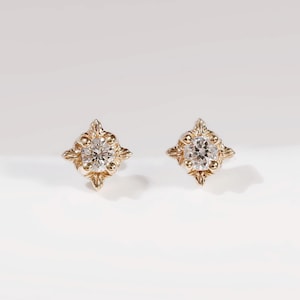 May include: A pair of gold stud earrings, each featuring a central round clear gemstone set within a four-pointed star-shaped frame with textured, beaded detailing along the edges, creating a classic and symmetrical appearance for the ears.