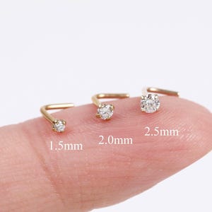 May include: Three gold L-shaped nose studs with clear round stones, labeled with sizes 1.5mm, 2.0mm, and 2.5mm. Each stud features a prong setting holding a single sparkling stone, showcasing the variation in size for these jewelry pieces.