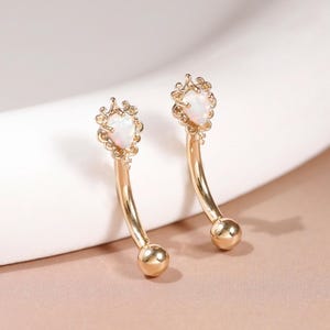 Pode incluir: Um par de brincos curvos de ouro com uma pedra de opala em forma de lágrima no topo e uma pequena esfera de ouro na base. A opala está cravada numa moldura dourada delicada e decorativa, criando um aspeto refinado e polido.