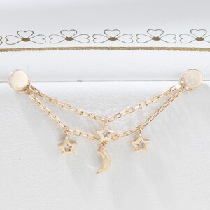 May include: A gold double-chain earring featuring two small star charms and a central crescent moon charm. Each star charm is accented with a tiny clear gemstone. The earring is designed with two flat circular posts at the ends of the chains.