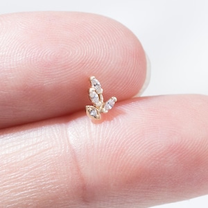 Op de afbeelding: Een kleine, goudkleurige oorbel met een bladachtig ontwerp. De oorbel heeft drie heldere, sprankelende edelstenen. De sieraden worden op een vinger tegen een witte achtergrond getoond. De oorbel is een delicaat sieraad.