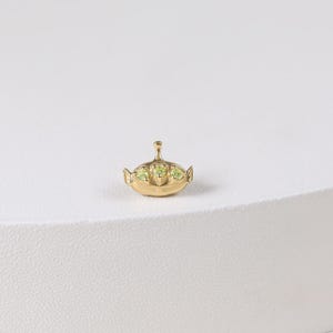 May include: A small, gold-colored charm in the shape of a Toy Story alien. The charm has three green gem-like eyes and a small antenna. The charm is set against a white background.