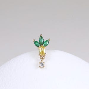 May include: A gold-toned earring with a pineapple design. The top features three green marquise-cut stones and a yellow oval-cut stone. Below, a series of smaller stones and a dangling clear stone complete the design. The earring is set against a white background.