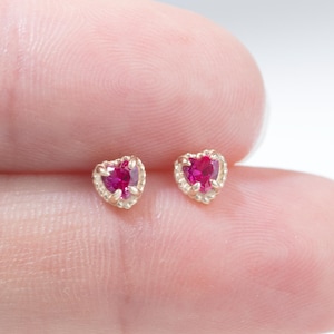 May include: A pair of heart-shaped stud earrings. Each earring has a vibrant red gemstone set in a gold-coloured frame. The earrings are small and delicate, suitable for everyday wear.