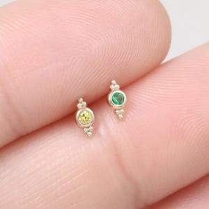 May include: Two small gold earrings, each with a central gemstone. One earring features a yellow gemstone, the other a green gemstone. The earrings have a decorative design with small details. The earrings are shown on a light background.