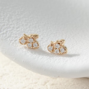 May include: A pair of gold-colored stud earrings. Each earring features a small, stylized design with three clear, round gemstones. The earrings are set against a white, textured surface.