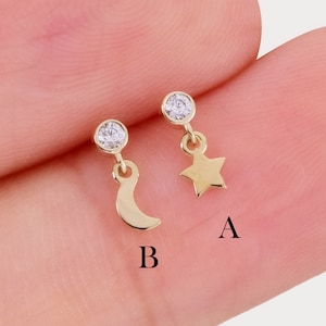Peut inclure: Deux boucles d'oreilles à tige dorées avec une petite pierre claire sur le dessus. L'une porte une breloque étoile étiquetée A, l'autre une breloque croissant de lune étiquetée B. Les boucles d'oreilles sont présentées sur un fond couleur peau.