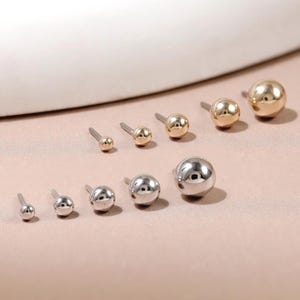 May include: A collection of ten spherical stud earrings arranged in two rows. The top row features five gold-toned ball studs of increasing size, while the bottom row displays five silver-toned ball studs, also arranged in order of increasing size.
