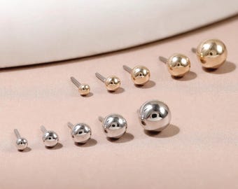 Può includere: Una collezione di dieci orecchini a lobo sferici disposti su due file. La fila superiore presenta cinque orecchini a sfera color oro di dimensioni crescenti, mentre la fila inferiore mostra cinque orecchini a sfera color argento, anch'essi disposti in ordine di dimensione crescente.