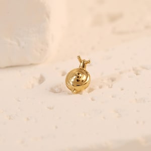 Può includere: Un piccolo ciondolo a forma di lumaca color oro. Il ciondolo presenta un design a conchiglia a spirale e delicate antenne. La lumaca è posizionata su una superficie strutturata bianco sporco, con uno sfondo morbido e neutro.