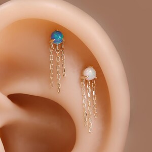 May include: Close-up of two gold earrings with a dangling chain design. One earring features a blue gemstone, and the other has a white gemstone. The earrings are displayed on an ear, showcasing their delicate design.