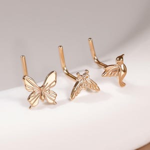 May include: Three gold-toned nose studs featuring distinct shapes: a butterfly, a moth, and a hummingbird. Each piece is crafted with an L-shaped post for secure placement, showcasing detailed metalwork and a polished finish on each animal design.