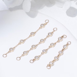 May include: Four gold-toned chain connectors, each featuring a series of clear, round gemstones. The connectors are arranged on a white surface, with a soft, blurred white floral arrangement in the background. The connectors are delicate and elegant.