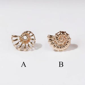 May include: Two gold-toned metal stud earrings shaped like ammonite fossils. Option A features an open, wire-like spiral structure, while option B displays a solid, textured, and detailed shell design. Both pieces are labelled with letters A and B.