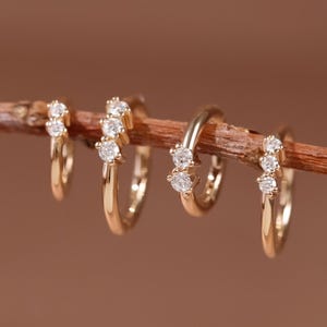 May include: Four gold hoop earrings are arranged in a row on a thin, brown wooden twig. Each earring features a small, circular band made of polished gold metal, accented with a cluster of three tiny, clear, round-cut gemstones set along the front.