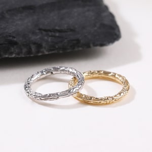 Könnte beinhalten: Zwei strukturierte Ringe, einer silberfarben und einer goldfarben, liegen auf einer weißen Oberfläche neben einem dunklen, strukturierten Stein. Die Ringe haben ein detailliertes, organisches Muster, das auf ein handwerkliches Design hindeutet. Der goldene Ring hat einen warmen, hellen Ton.