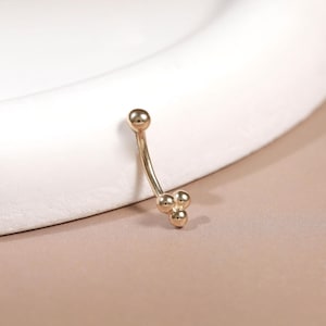 May include: A curved, gold-colored belly button ring. The jewelry features a small ball at the top and three smaller balls clustered together at the bottom. The ring is set against a neutral background.