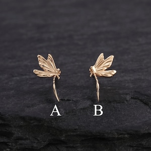 May include: Two gold-colored dragonfly earrings, each with delicate wing details and a slender body. The earrings are displayed on a dark, textured surface, with the letters "A" and "B" beneath each earring.