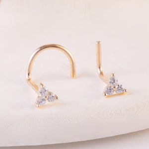 May include: Two gold-toned nose studs featuring a triangular cluster of three small, clear, sparkling stones. One stud has a curved post, while the other has an L-shaped post. The jewelry pieces are metallic gold in color with clear stones.