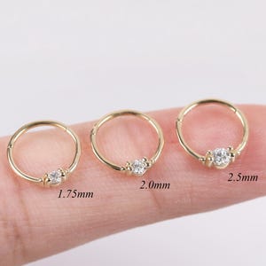 May include: Three gold-colored nose rings with a small, clear gemstone in the center. The rings are different sizes, with measurements of 1.75mm, 2.0mm, and 2.5mm. The rings are displayed on a finger against a white background.