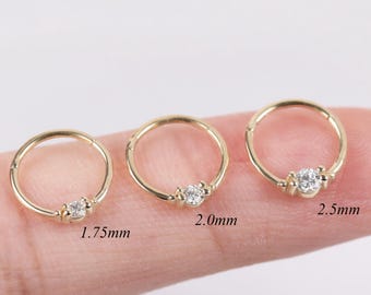 May include: Three gold-colored nose rings with a small, clear gemstone in the center. The rings are different sizes, with measurements of 1.75mm, 2.0mm, and 2.5mm. The rings are displayed on a finger against a white background.