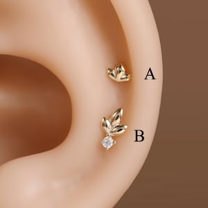 Könnte beinhalten: Zwei goldene Ohrstecker mit den Bezeichnungen A und B. Stecker A hat die Form einer kleinen Blume mit drei Blütenblättern. Stecker B zeigt ein dreiblättriges Design mit einem klaren, runden Stein an der Basis, platziert am Ohrknorpel.