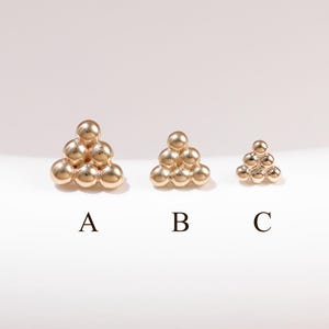 May include: Three gold-toned stud earrings arranged in a triangular cluster of six small spheres. The earrings are labeled A, B, and C, showing a progression in size from largest to smallest, each featuring a polished, reflective metallic finish.