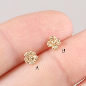 May include: Two small, gold-colored flower-shaped stud earrings. The earrings are positioned on a light background. The earrings have a detailed, textured design, with visible petal-like structures. The earrings are small, delicate, and likely for pierced ears.