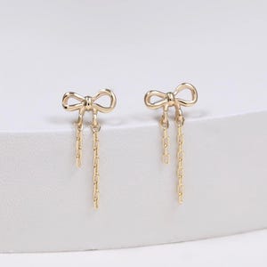 Peut inclure: Une paire de boucles d'oreilles dorées avec un motif de nœud. Chaque boucle d'oreille comporte deux fines chaînes dorées de longueurs différentes suspendues sous le nœud, créant un effet pendant subtil avec une finition métallique polie.