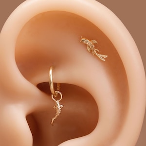 May include: A gold ear piercing set consisting of a small hoop earring with a dangling koi fish charm and a separate flat-back stud earring shaped like a koi fish, both featuring detailed scales and fins, positioned in the upper ear cartilage.