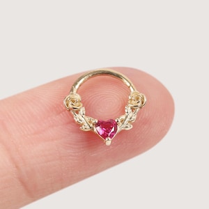 Op de afbeelding: Een goudkleurige neusring met een roze, hartvormige edelsteen in het midden. De ring is versierd met bloemen- en bladerdetails. De sieraden worden door een vinger tegen een neutrale achtergrond gehouden.