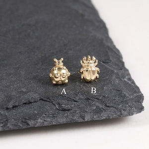 Könnte beinhalten: Zwei kleine, goldfarbene Insekten-förmige Anhänger, beschriftet mit A und B, liegen auf einer dunkelgrauen Oberfläche. Anhänger A ähnelt einem Marienkäfer, während Anhänger B ein Skarabäus-Käfer ist. Die Anhänger sind mit strukturierten Oberflächen versehen und sind wahrscheinlich Schmuckkomponenten.