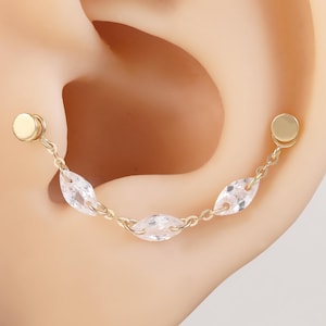 Op de afbeelding: Een goudkleurige kettingoorbel met drie heldere, marquise-geslepen stenen die tussen twee platte, ronde knopjes hangen. De fijne metalen ketting verbindt de knopjes en creëert een gedrapeerd effect over het oorkraakbeen.