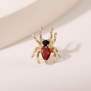 May include: A gold-toned spider-shaped stud earring featuring a small black circular stone for the head and a larger pear-shaped red gemstone for the abdomen. The spider has eight curved legs extending from the central body structure.