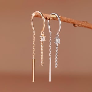 May include: Two threader earrings hanging from a thin twig. One earring is rose gold and the other is silver. Each earring features a small clear gemstone set in a prong setting, connected to a delicate chain that ends in a smooth metal bar.