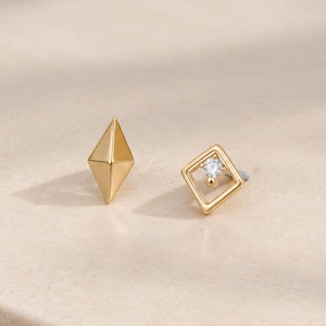 May include: A pair of gold stud earrings featuring two distinct shapes. One earring is a solid, three-dimensional diamond-shaped stud with faceted edges. The other earring is a square frame stud with a small, clear, round gemstone set at the center.