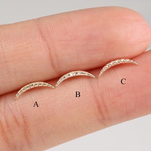 Op de afbeelding: Drie goudkleurige, halvemaanvormige oorbellen, gelabeld met A, B en C. Elke oorbel heeft een ander ontwerp. Oorbel A heeft kleine, gelijkmatig verdeelde details. Oorbel B heeft een glad oppervlak met een paar details. Oorbel C is bezet met kleine, sprankelende steentjes.