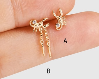 May include: Two gold-coloured scorpion earrings. One earring is a stud with a small clear gem. The other earring has a scorpion design with dangling chains and two gold-coloured bars. The earrings are on a light grey background.