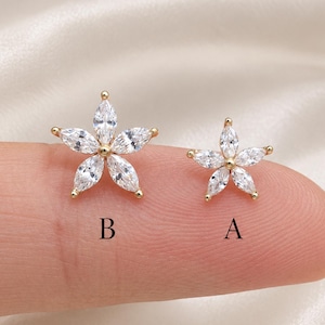 May include: Two gold-toned stud earrings shaped like five-petaled flowers, each featuring marquise-cut clear cubic zirconia stones. The earring labeled B is larger than the earring labeled A. Both pieces have a central gold-toned metal setting.