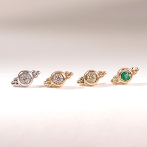 May include: Four stud earrings arranged in a horizontal row. Each earring features a central round gemstone set in a bezel, flanked by three small decorative metal spheres on each side. The stones include clear, light yellow, and green gems.