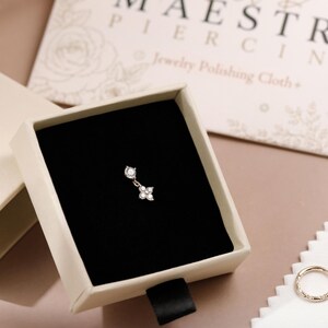 May include: A small gold piercing charm with two clear stones, one round and one flower-shaped, resting inside a cream-colored jewelry box with a black velvet insert. The box is labeled Gold Maestro, and a matching gold ring sits nearby.
