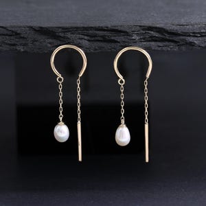 May include: A pair of gold threader earrings featuring a curved arch top, a delicate chain segment ending in a small white teardrop-shaped pearl, and a straight metal bar on the opposite side of the arch, creating a balanced and symmetrical design.