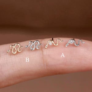 May include: Four small snake-shaped stud earrings arranged in a row. Two are gold-toned and two are silver-toned. The two earrings on the right are marked with the letters A and B, and each features a small, clear, round-cut gemstone set in the head.